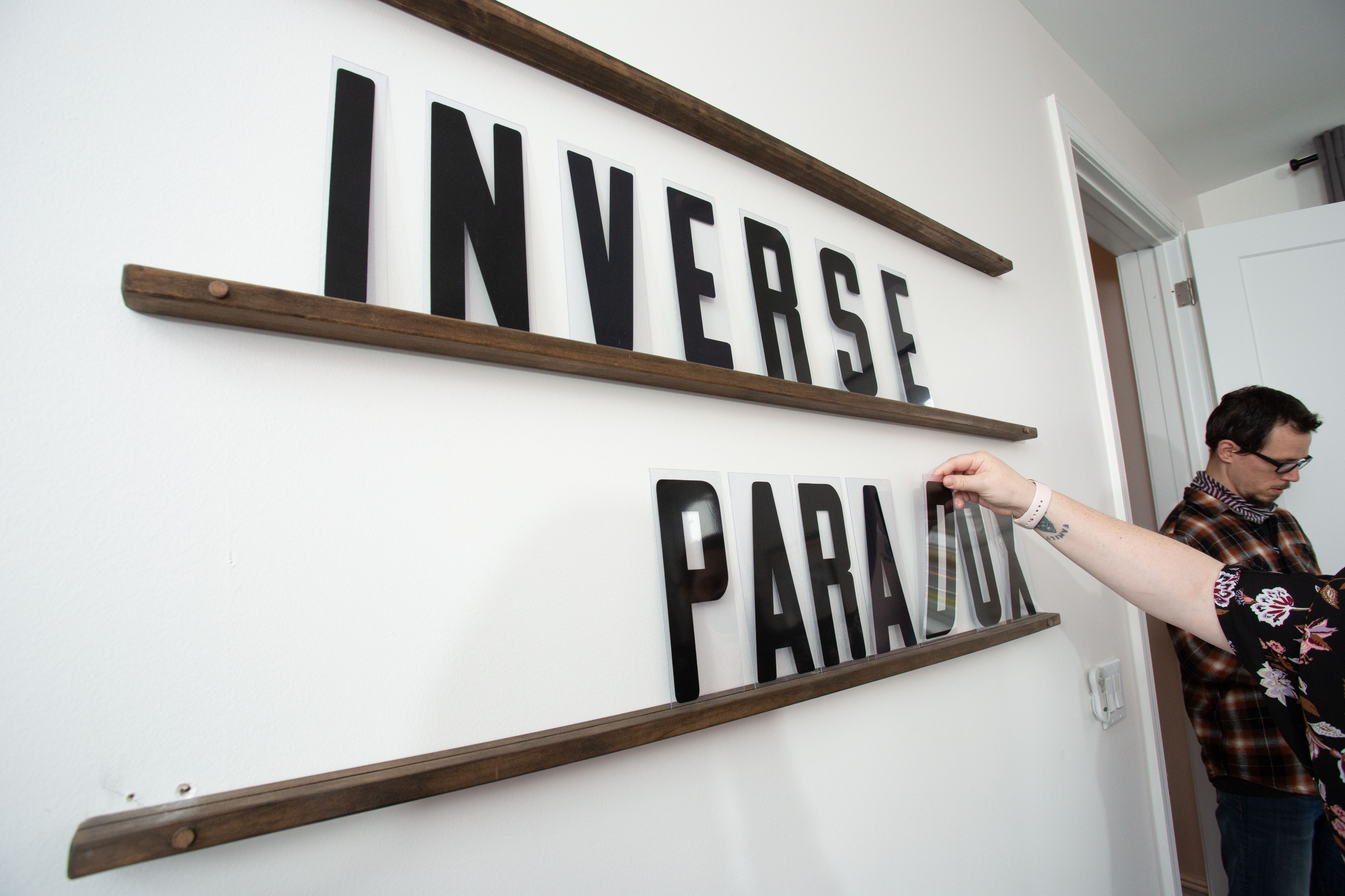 A wall with three thin shelves and letters that spell out Inverse Paradox two people stand nearby one looking down and the other holding the letter 'd' in their hand
